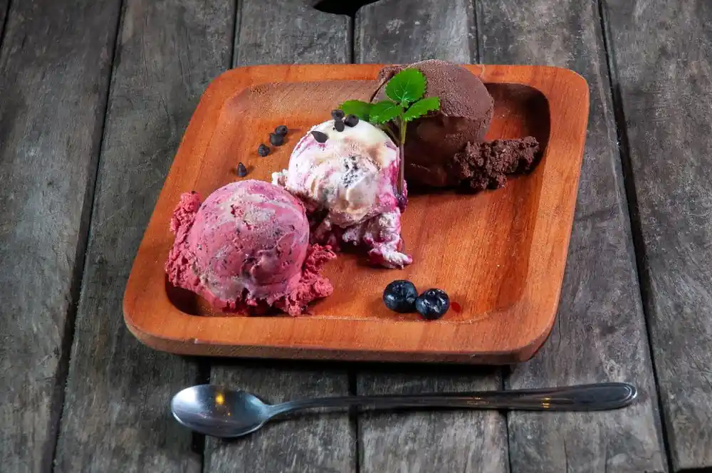 Three artisanal ice cream scoops served on a wooden plate with fresh berries and dark chocolate